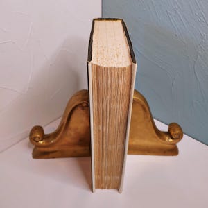 May include: A vintage book with aged, cream-colored pages and a black spine, standing between two ornate, gold-colored bookends. The bookends have a curved design with a scrolled detail.