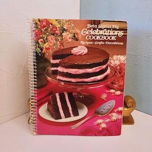 May include: A spiral-bound cookbook titled "Beta Sigma Phi Celebrations COOKBOOK" with a chocolate cake on the cover. The cake has pink frosting layers and a slice is on a plate. The cover also features text: Recipes, Crafts, Decorations.