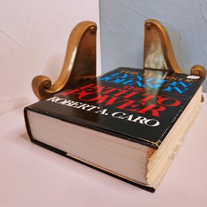 May include: A hardcover book titled "The Years of Lyndon Johnson: The Path to Power" by Robert A. Caro, held between two ornate, gold-colored bookends. The book has a black cover with red and blue text.