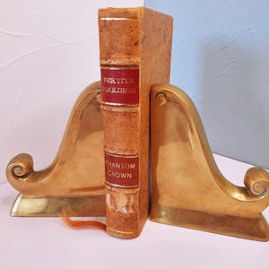 May include: A pair of gold metal bookends with a book titled "Phantom Crown" by "Bertile Harding" in between them.