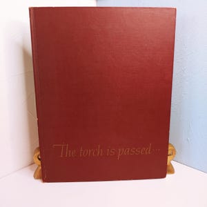 May include: A burgundy-colored book with the gold text "The torch is passed..." on the cover. The book is propped up by a gold-colored book stand. The book has a textured cover.
