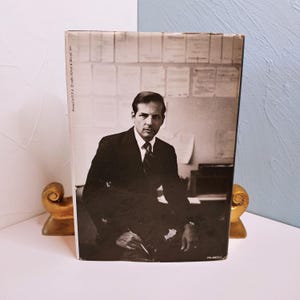 May include: A vintage book cover featuring a black and white photograph of a man in a suit. The man is seated, looking directly at the viewer. The book is propped up by a gold-colored decorative bookend.