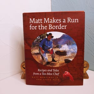 May include: A cookbook titled "Matt Makes a Run for the Border" with a photo of a man cooking outdoors. The cover also reads "Recipes and Tales from a Tex-Mex Chef" with the authors' names.