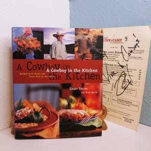 May include: A cookbook titled "A Cowboy in the Kitchen" with images of food and a cowboy. The book is open, revealing a menu with handwritten signatures. The cover features a man in a cowboy hat and various food photos.