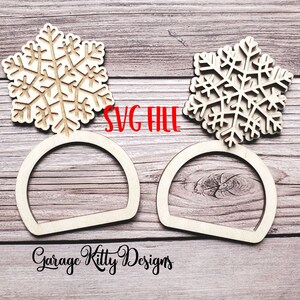 May include: Two wooden snowflake ornaments with a semi-circular base. The snowflakes are intricately cut with detailed patterns. The text "SVG FILE" is in red. The words "Garage Kitty Designs" are in black script.
