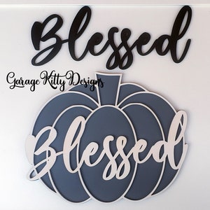 May include: Wooden sign with the word "Blessed" in a cursive font. A pumpkin-shaped sign in shades of gray and white also features the word "Blessed". The text "Garage Kitty Designs" is also visible.