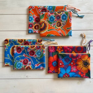 May include: Five colorful zippered pouches with floral patterns. The pouches are made of fabric and have a variety of colors, including blue, orange, red, yellow, and green. The pouches are arranged in a row on a white wooden surface.