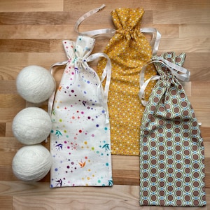 May include: Three white yarn balls next to three drawstring bags. The bags have different patterns: a white bag with colorful birds and stars, a yellow bag with white polka dots, and a green bag with brown circles.