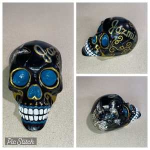 May include: A black skull figurine with teal eye sockets and gold accents. The skull has white teeth and the name "Jazmin" in gold script. A cat is painted on the side of the skull.