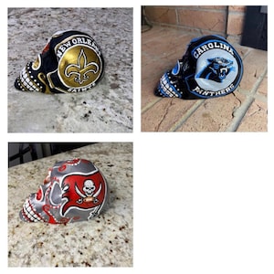 Könnte beinhalten: Drei dekorative Totenkopf-Figuren. Eine ist schwarz und goldfarben mit dem Logo der New Orleans Saints. Eine andere ist blau und schwarz mit dem Logo der Carolina Panthers. Die dritte ist grau, rot und weiß mit dem Logo der Tampa Bay Buccaneers.