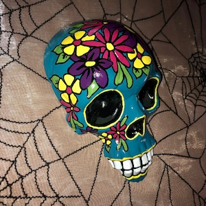 Könnte beinhalten: Ein türkisfarbener Keramikschädel, verziert mit bunten Blumen in Rosa-, Gelb- und Lilatönen. Der Schädel hat schwarze Augenhöhlen und ein weißes Grinsen. Der Schädel liegt auf einer braunen Oberfläche mit einem schwarzen Spinnennetz-Design.