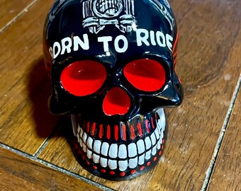 Op de afbeelding: Zwart schedelvormig decoratief item met rode oogkassen en mond. De schedel heeft witte tanden en de woorden "BORN TO RIDE" in witte letters. Een motorfietsafbeelding staat boven de tekst.