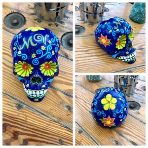 Könnte beinhalten: Eine Sammlung von drei dekorativen Totenkopf-Schädeln zum Tag der Toten. Die Schädel sind in leuchtenden Farben bemalt und weisen komplizierte florale Designs und Muster auf. Die Hauptfarbe ist ein tiefes Blau mit Akzenten in Gelb, Orange und Grün.