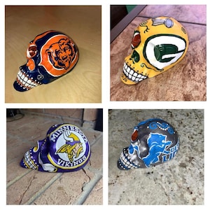 May include: Four hand-painted ceramic skulls, each featuring a different NFL team logo. The skulls are painted in team colors, including orange and navy for the Chicago Bears, green and yellow for the Green Bay Packers, purple and gold for the Minnesota Vikings, and blue and silver for the Detroit Lions.