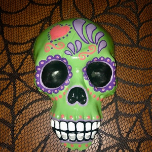 May include: A Day of the Dead-themed skull decoration. The skull is painted light green with black eye sockets and a black nose. It has purple and pink floral designs and white teeth. The skull is on a brown surface with a black web design.