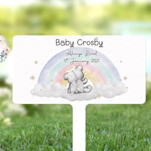 Può includere: Un paletto di legno bianco con un arcobaleno e un orsacchiotto con le ali d'angelo. Il paletto riporta la scritta "Baby Crosby Always Loved 17th January 2021".