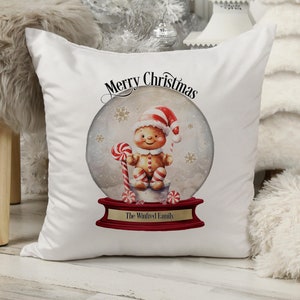 Pode incluir: Travesseiro branco com um design de bola de neve que apresenta um homem de gengibre com um chapéu vermelho segurando um bastão de caramelo. O texto "Merry Christmas" está acima da bola de neve e "The Winifred Family" está abaixo da bola de neve.