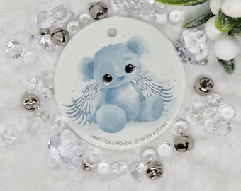 May include: A white ceramic ornament with a blue teddy bear with wings. The teddy bear has a sweet expression and is sitting with its wings spread out. The ornament has the text "Carried for a moment, loved for a lifetime" on the bottom.