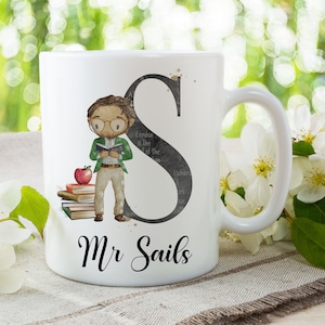 Può includere: Tazza in ceramica bianca con un'illustrazione a cartoni animati di un insegnante che legge un libro, con una mela e libri. La tazza ha il nome "Mr Sails" e una grande lettera "S". Lo sfondo è sfocato con fiori bianchi.