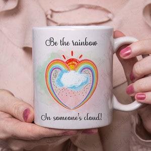Puede incluir: Taza de cerámica blanca con un diseño de corazón arcoíris y el texto "Sé el arcoíris en la nube de alguien!"