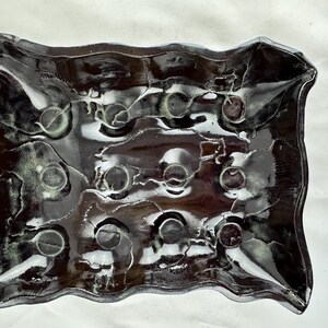 Puede incluir: Un plato de cerámica negro con un borde ondulado y una superficie texturizada. El plato tiene una serie de hendiduras circulares.