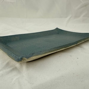 Puede incluir: Una bandeja rectangular de cerámica con un esmalte azul claro. La bandeja tiene un borde ligeramente elevado y una superficie texturizada.