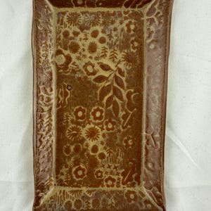 May include: A rectangular ceramic tray with a reddish-brown glaze and a lighter tan floral pattern embossed across the surface. The tray features slightly raised, flared edges and a flat base, displaying a consistent botanical motif throughout.