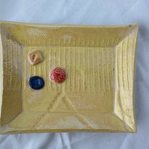May include: A yellow ceramic rectangular tray with a raised border. The tray has a textured surface and three decorative accents: a red button, a blue button, and a small yellow square.
