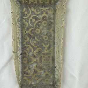 May include: A rectangular ceramic tray with a light green and grey mottled glaze. The surface features a repeating intricate geometric pattern in a lighter shade, with textured, uneven edges and a slightly flared top rim design.