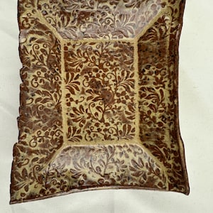 Può includere: Un piatto quadrato decorativo in ceramica con un motivo floreale marrone e beige. Il piatto ha una superficie leggermente rialzata, strutturata e un bordo smerlato. Il motivo floreale è ripetuto su tutto il piatto.