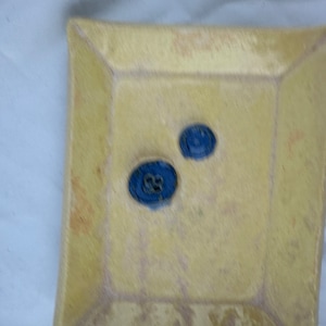 May include: A square ceramic dish with a light yellow glaze. The dish has two blue circular stamps on the surface.