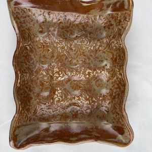 Puede incluir: Un plato cuadrado de cerámica con un borde ondulado y un glaseado marrón. El plato tiene un diseño en relieve de hojas y enredaderas en espiral en un color marrón más claro.