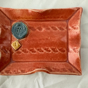 May include: A small, rectangular ceramic dish with a red glaze and a wavy edge. The dish has a decorative rope design and two small, round ceramic embellishments, one blue and one yellow.