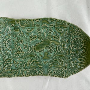 May include: A green ceramic serving dish with a raised, embossed floral pattern. The dish has an irregular, organic shape.