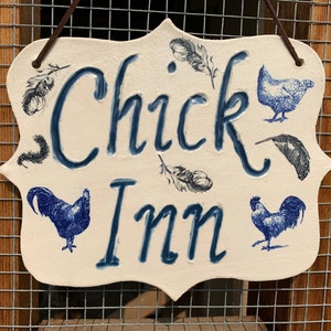 以下が含まれることがあります： クリーム色の装飾的な看板で、濃い青色で「Chick Inn」の文字が書かれています。鶏と羽のイラストが描かれています。茶色のコードで吊り下げられており、金網の背景に配置されています。