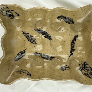 Puede incluir: Porta huevos de cerámica color beige con borde festoneado. El soporte presenta diseños de plumas negras e indentaciones circulares para huevos. La superficie moteada añade un toque rústico a este accesorio de cocina.