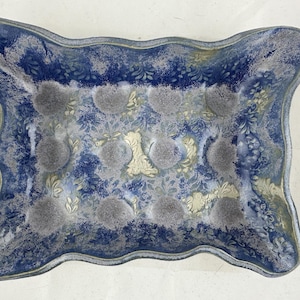 Puede incluir: Un plato de servir de cerámica azul con un borde ondulado y un diseño decorativo de patrones florales blancos y dorados. El plato tiene un esmalte moteado y un aspecto rústico.