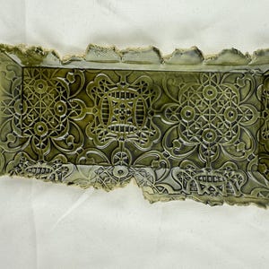 May include: A rectangular ceramic tray with a glossy olive green glaze. The surface features a repeating embossed geometric pattern with intricate lattice and floral-inspired motifs. The edges of the tray have a rough, uneven, and textured finish.