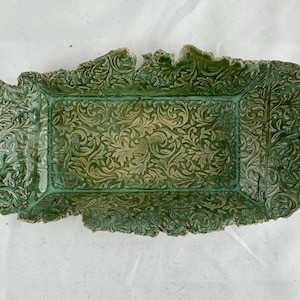 Pode incluir: Uma bandeja de servir de cerâmica verde com um design floral em relevo. A bandeja tem uma forma irregular com uma borda ligeiramente elevada.