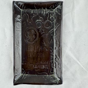 Op de afbeelding: Een donkerbruine keramische dienblad met een reliëfdesign met Harry Potter-themabeelden, waaronder het Hogwarts-wapen, brillen, een bezem en het nummer 9 3/4. De dienblad bevat ook de tekst "LevioSA LevioSA" en "Mischief Managed".