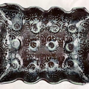 Puede incluir: Un plato de servir de cerámica marrón oscuro con un borde ondulado y un diseño intrincado en relieve en un tono de verde más claro. El plato es perfecto para servir aperitivos o aperitivos.