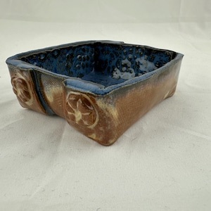 May include: A square ceramic bowl with a textured brown exterior and a deep blue interior. The outer sides feature raised circular floral motifs, while the inner surface displays a mottled, glossy blue glaze with a distinct pitted texture.
