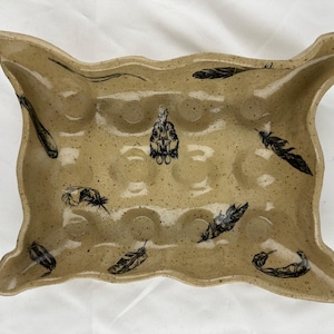 Puede incluir: Portahuevos de cerámica beige con borde ondulado, superficie moteada e ilustraciones de plumas y calaveras negras. El soporte tiene 12 hendiduras circulares para huevos.