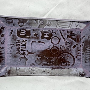 May include: A rectangular ceramic dish with a purple glaze and a white design. The design features various Harry Potter themed symbols, including a wand, glasses, a star, and the words "Swish & Flick".