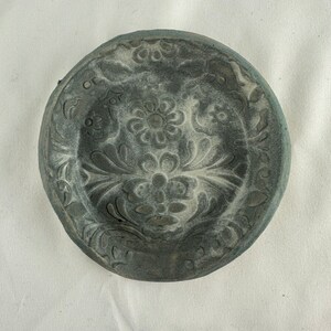 May include: A gray ceramic dish with a raised floral design. The dish has a slightly rounded shape and a smooth, matte finish.