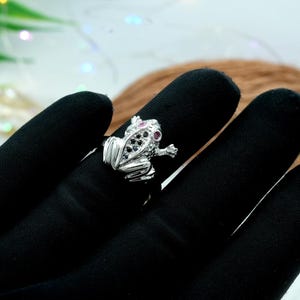 Puede incluir: Un anillo de plata con forma de rana con piedras preciosas negras y rosas para los ojos. El anillo presenta detalles plateados y se encuentra sobre un fondo negro.