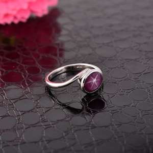 May include: A silver ring with a pink star-shaped gemstone. The ring is on a black textured surface.
