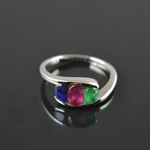 Peut inclure: Une bague en argent avec trois pierres précieuses : un saphir bleu, un rubis rouge et une émeraude verte. La bague est sertie sur une bande incurvée.