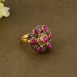 May include: A gold ring featuring a central cluster design with a round cabochon ruby surrounded by smaller oval cabochon rubies, a ring of tiny green stones, and a dark metal base encrusted with numerous small clear white stones.