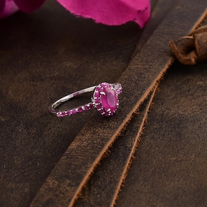 Puede incluir: Un anillo de plata con una gran piedra de rubí ovalada rodeada de piedras de rubí más pequeñas. El anillo está sobre una superficie de cuero marrón.